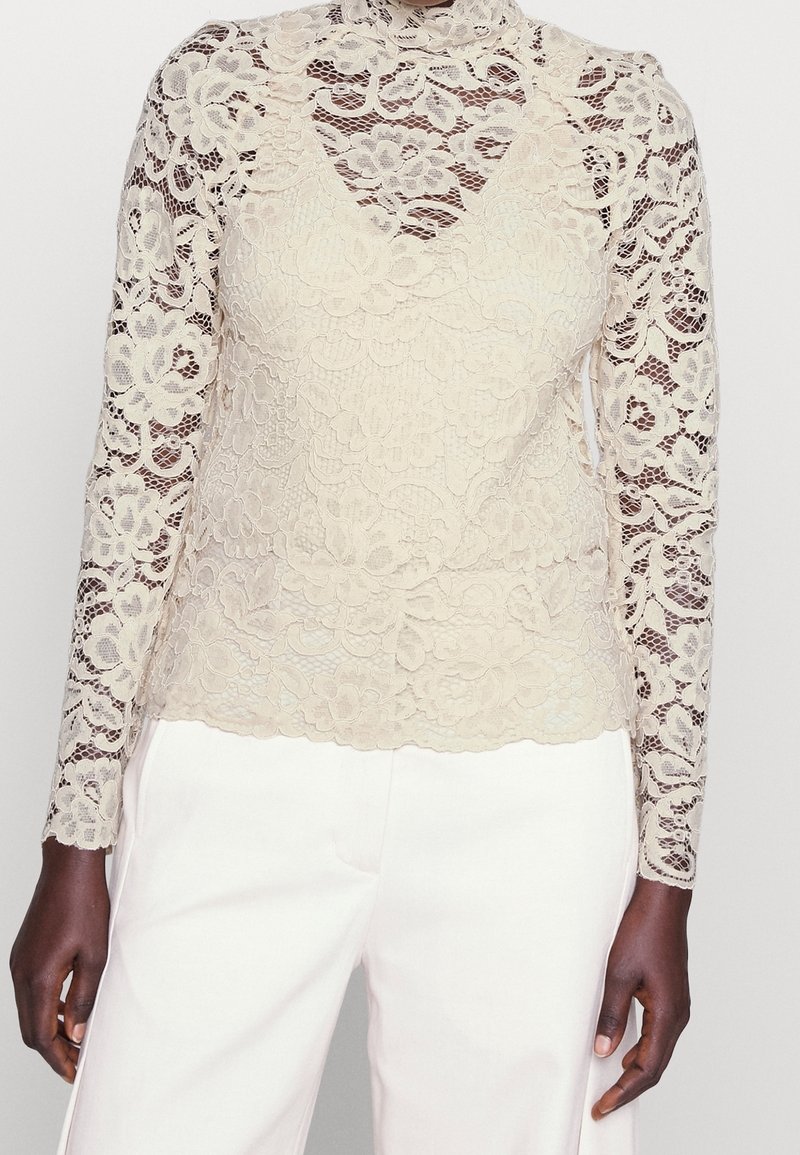 Beige lace top with long sleeves, floral pattern, high neckline, and sheer detailing. Paired with white pants. Textured finish throughout.