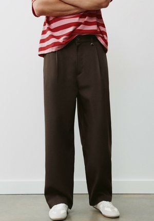 Personne portant un pantalon marron à jambes larges, des baskets blanches et une chemise rayée rouge et rose, les bras croisés.