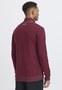 Burgunderfarbener Strickpullover mit hohem Kragen, strukturiertem Gewebe, kontrastierenden Rippakzenten und einem kleinen Etikett am hinteren Kragen.