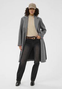 Manteau en laine gris, col roulé beige, jeans noirs, ceinture marron avec boucle en or, bottes noires à talons, et une casquette beige. Cheveux bouclés visibles.