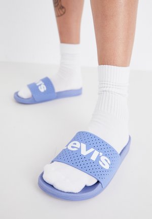 Sandalias de diapositivas azul claro con una correa texturizada que presenta el logo blanco de "Levi's". Usadas con calcetines blancos hasta la mitad de la pierna, mostrando un estilo relajado.