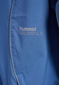 Blå techwear-tyg har ett strukturerat rutmönster. Logotypen "hummel" och ytterligare text är tryckta i ljusgrått.