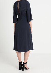 Námořnické šaty se stáhnutým pasem, rozšířenými rukávy a detaily ve tvaru klíčové dírky na zádech. Hladká textura a délka ke kolenům doplňují černé sandály na tenkých podpatcích.