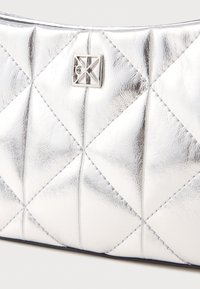 Ezüst, steppelt estélyi tasak fényes, texturált felülettel. Kis, geometrikus fém logó díszítéssel rendelkezik. Téglalap alakú, varrásos részletekkel.