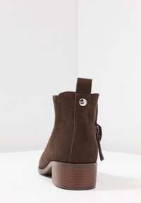 Hnědé semišové kotníkové boty s nízkým podpatkem, bočním zipem a poutkem na zapnutí, které má malou kovovou logovou knoflíkovou sponu.