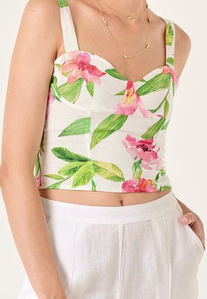 Woman wearing a white floral crop top with pink flowers and green leaves, paired with high-waisted white pants and a delicate gold necklace.