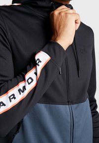 Sort og grå zip-up hættetrøje med et kontrasterende hvidt og orange bånd med "ARMOUR" trykt på ærmet. Glat stof og justerbar hætte.