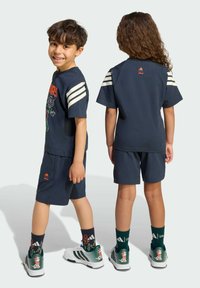 T-shirt blu navy a maniche corte con strisce bianche, caratterizzata da una grafica colorata Disney, abbinata a pantaloni corti coordinati e sneakers verdi.