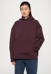Felpa con cappuccio bordeaux realizzata in tessuto morbido, con tasca frontale, vestibilità comoda e polsini a costine, abbinata a jeans blu.