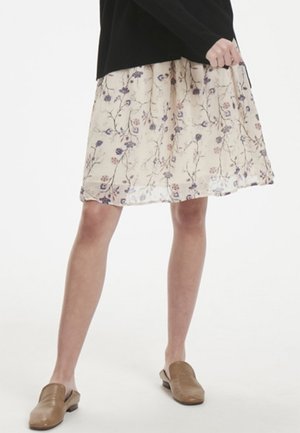 Jupe midi à imprimé floral en tissu beige doux avec un design fluide, ornée de fleurs violettes et roses, accompagnée de chaussures marron à enfiler.