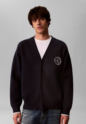 Jeune homme portant un cardigan noir boutonné avec un logo Calvin Klein et une chemise blanche, debout devant un fond gris uni.