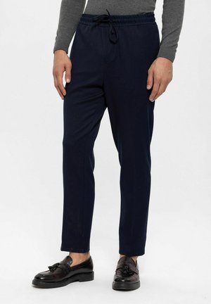 Pantalon bleu marine avec une ceinture à cordon, coupe droite, texture lisse, assorti à des mocassins noirs. Style minimaliste.