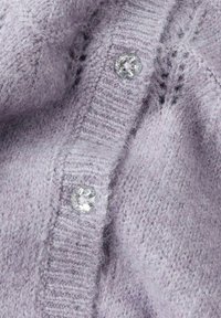 Lilla strikket cardigan med en teksturert stoff, ribbede kanter og klare dekorative knapper. Nærbilde av knapp- og søm detaljer.