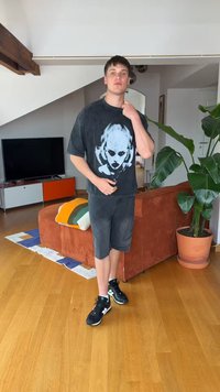 Νέος άντρας στέκεται σε ένα σαλόνι φορώντας μαύρο γραφικό μπλουζάκι, σκούρα σορτς, μαύρα αθλητικά παπούτσια, κοντά σε καφέ πορτοκαλί καναπέ και μεγάλο γλαστρωμένο φυτό.