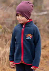 Marine fleecejakke med rød lynlås og kant, der har et legende patch-design. Blød tekstur, lange ærmer og høj krave. Brun hue.