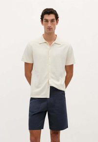 Polo shirt crème à manches courtes avec col, présentant un devant boutonné et une coupe décontractée, associé à un short bleu marine foncé. Texture lisse.