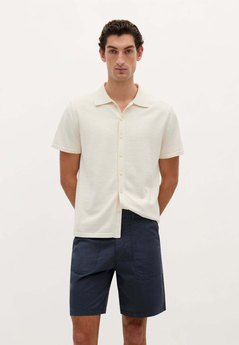 Polo shirt crème à manches courtes avec col, présentant un devant boutonné et une coupe décontractée, associé à un short bleu marine foncé. Texture lisse.