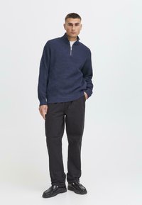 Pullover en tricot bleu marine avec un zip quart et un motif texturé, associé à un pantalon noir à coupe droite et des chaussures noires. Design minimaliste.
