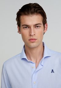 Chemise à carreaux bleu clair avec un col boutonné et un logo de crâne marine sur la poitrine gauche. Tissu doux avec une texture lisse.