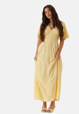 Femme aux longs cheveux foncés portant une robe maxi vichy jaune avec des boutons et des talons ouverts beige, debout devant un fond blanc.
