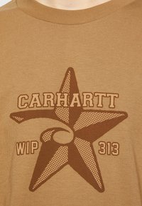 Ruskea puuvillapaita, jossa on suuri ruskea ja beige tähti graafinen kuvio, jossa on pistekuvio, sekä tekstit "CARHARTT WIP" ja "313" alla.