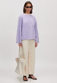 Lila lange mouwen shirt gecombineerd met romige wijde broek; houdt een romige tote bag vast; zwarte zonnebril; zwarte flip-flops, vlakke textuur.
