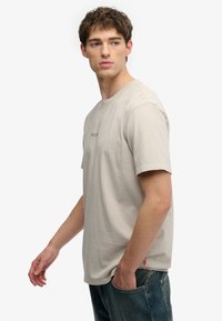 Lysebeige bomulds-T-shirt med rund hals, korte ærmer og subtilt broderet logo foran, kombineret med blå denimjeans.