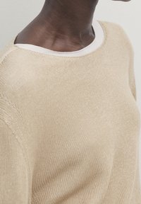 Pull en maille beige avec un col rond, présentant une texture lisse et un col intérieur blanc contrastant. Vue rapprochée de l'épaule.