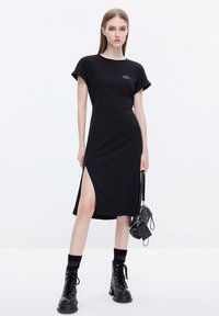 Vestido negro de camiseta con mangas cortas, aberturas laterales y una textura suave. Presenta un pequeño detalle gráfico en el pecho. Combinado con botas de combate negras.