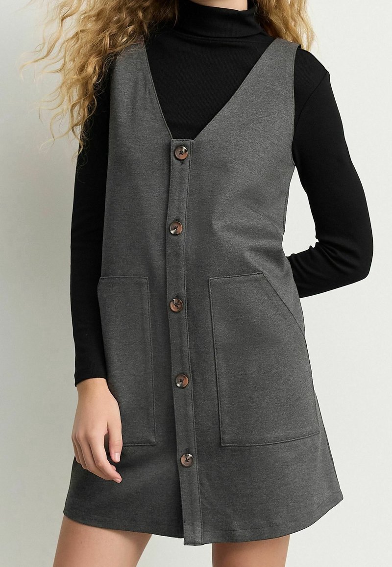 Femme portant une robe sans manches gris foncé à boutons sur le devant avec de grandes poches, par-dessus un haut noir à manches longues et col roulé.