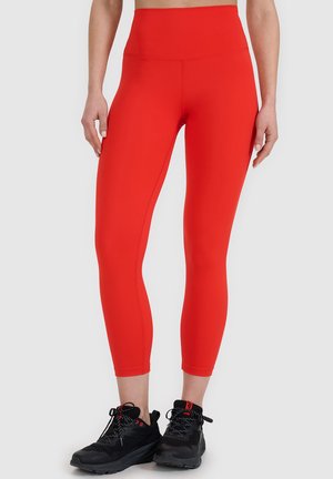 Osoba nosząca jasnoczerwone krótkie legginsy z wysokim stanem i czarne buty sportowe stoi na jednolitym tle.