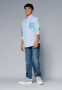 Camisa de mangas compridas riscada em azul claro com um bolso azul, combinada com jeans azuis e sapatilhas cinzas, contra um fundo cinza.
