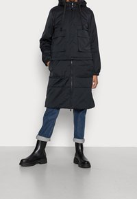 Czarna pikowana parka z zamkiem błyskawicznym, dwoma przednimi kieszeniami, elastycznymi mankietami i kapturem, zestawiona z podwiniętymi niebieskimi dżinsami i czarnymi butami Chelsea.