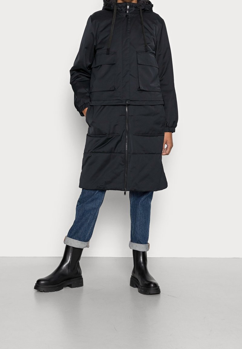 Czarna pikowana parka z zamkiem błyskawicznym, dwoma przednimi kieszeniami, elastycznymi mankietami i kapturem, zestawiona z podwiniętymi niebieskimi dżinsami i czarnymi butami Chelsea.