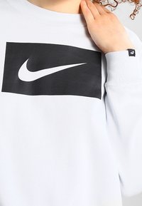 Světle modrá mikina s černým obdélníkovým logem Nike na přední straně. Texturovaná tkanina s žebrovanými manžetami a malou značkovou cedulkou na rukávu.