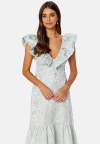 Světle mátově zelené šaty s květinovým texturovaným vzorem, hlubokým V-výstřihem, volánkovými rukávy a rozšířeným lemem.