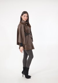 Bruine neplederen poncho met een hoge kraag en een ceintuur in de taille, gecombineerd met donkergrijze skinny jeans en zwarte enkellaarzen.