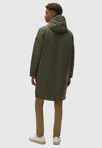 Gabardina verde oliva larga con capucha, hecha de un material suave. Presenta un corte recto y se combina con pantalones color tostado y zapatillas blancas.