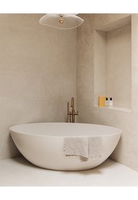 Vasca da bagno ovale freestanding in bianco opaco con un asciugamano grigio chiaro adagiato sul bordo, abbinata a un rubinetto dorato contro una parete beige testurizzata.