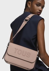 Borsa a spalla in pelle rosa con logo "TOUS" in rilievo, forma rettangolare, tracolla regolabile e dettagli in metallo argentato. Texture liscia.
