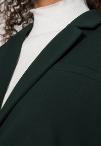 Tmavě zelený blazer s klopami, hladkou texturou a minimálním detailním šitím, nošený přes bílý žebrovaný rolák.