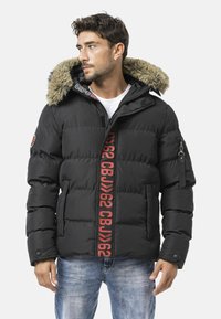Schwarze Daunenjacke mit einem Kapuzenfutter aus Fell, mit einem auffälligen roten Logostreifen in der Mitte und seitlichen Reißverschlusstaschen. Strukturierte, gesteppte Optik.
