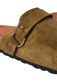 Chaussure slip-on en daim marron avec une large sangle, un anneau en métal et deux clous décoratifs. Surface texturée et semelle en caoutchouc noir contrastante.