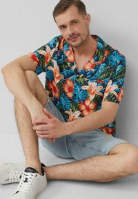 Camicia hawaiana a maniche corte con un motivo floreale e di foglie colorato su uno sfondo scuro. Pantaloni corti in denim azzurro chiaro e sneaker bianche.