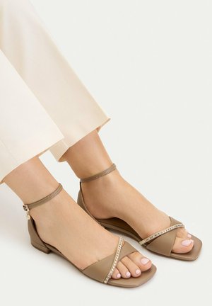Fødder iført beige åbne sandaler med ankelstropper og rhinstenpynt, kombineret med cremefarvede bukser mod en ensfarvet baggrund.