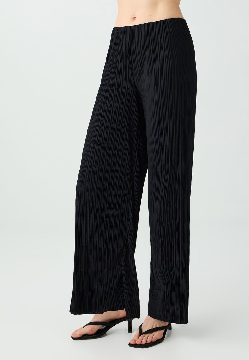 Pantalon noir plissé à coupe large porté avec des sandales noires à talons et bride entre les orteils, présenté de la taille aux pieds sur un fond clair.