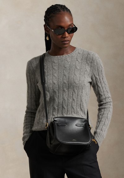 Femme portant des lunettes de soleil noires, un pull en maille torsadée gris, un pantalon noir et un sac bandoulière en cuir noir, debout devant un fond neutre.