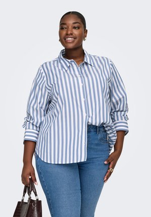 Lächelnde Frau, die ein blau-weiß gestreiftes Hemd und blaue Jeans trägt, hält eine dunkelbraune Handtasche mit kurzen Griffen.