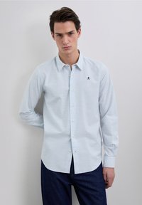 Lichtblauw overhemd met lange mouwen en een subtiel ruitpatroon, een button-down kraag en een klein zwart geborduurd schedellogo op de borst.