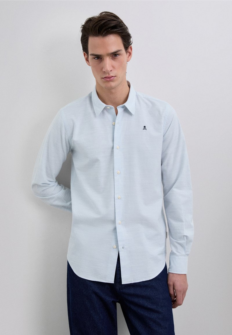 Lichtblauw overhemd met lange mouwen en een subtiel ruitpatroon, een button-down kraag en een klein zwart geborduurd schedellogo op de borst.